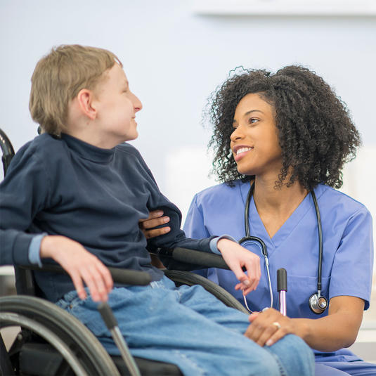 A nurse is helping a boy with cerebral palsy get into his wheelchair. They are both smiling and talking to each other. A nurse is helping a boy with cerebral palsy get into his wheelchair. They are both smiling and talking to each other.