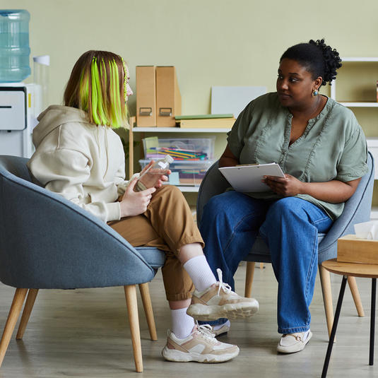 African female social worker talking to difficult teenage girl while they sitting on armchairs at office African female social worker talking to difficult teenage girl while they sitting on armchairs at office